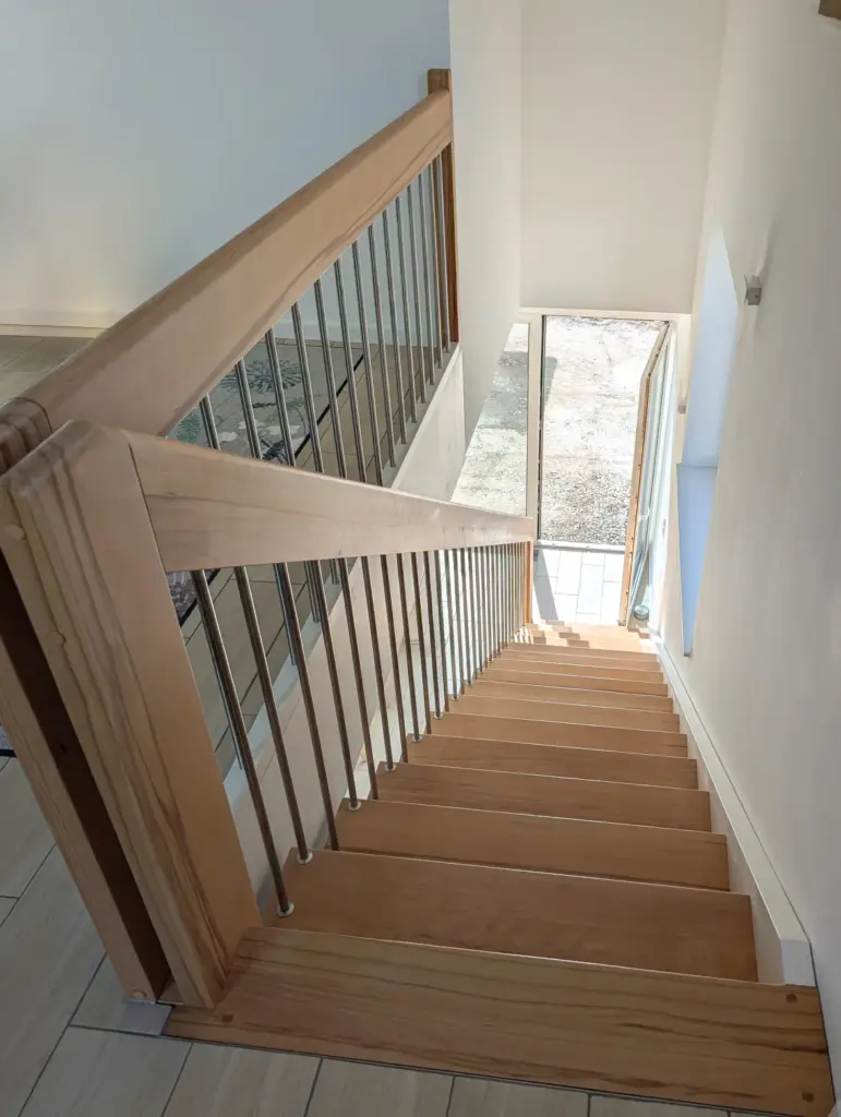 Eine Holztreppe mit Metallgeländer führt hinunter zu einer Glastür, die das Sonnenlicht hereinlässt und den Blick auf einen Innenhof und einen Außenbereich freigibt. Die Wände sind weiß und die Böden bestehen aus hellem Holz.