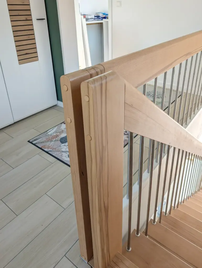 Holztreppe mit vertikalen Metallgeländerstäben, von oben gesehen. Der Handlauf und der Pfosten sind aus hellem Holz, und der Bereich darunter besteht aus Fliesen, einer Tür und einer Matte.
