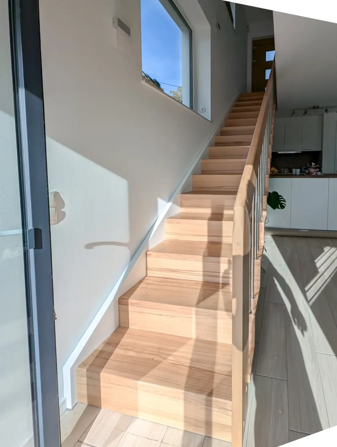 Eine Holztreppe mit einfachem Handlauf führt in einem hellen, modernen Wohnbereich mit großen Fenstern, hellen Holzböden und einem Blick auf den Küchenbereich im Hintergrund nach oben.
