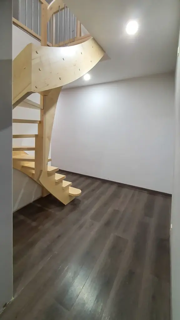 Die moderne Einrichtung besteht aus einem dunklen Holzfußboden, weißen Wänden und einer offenen Holztreppe mit geschwungenen Stufen, die in den zweiten Stock führt. Der Raum ist durch Deckenleuchten gut beleuchtet.