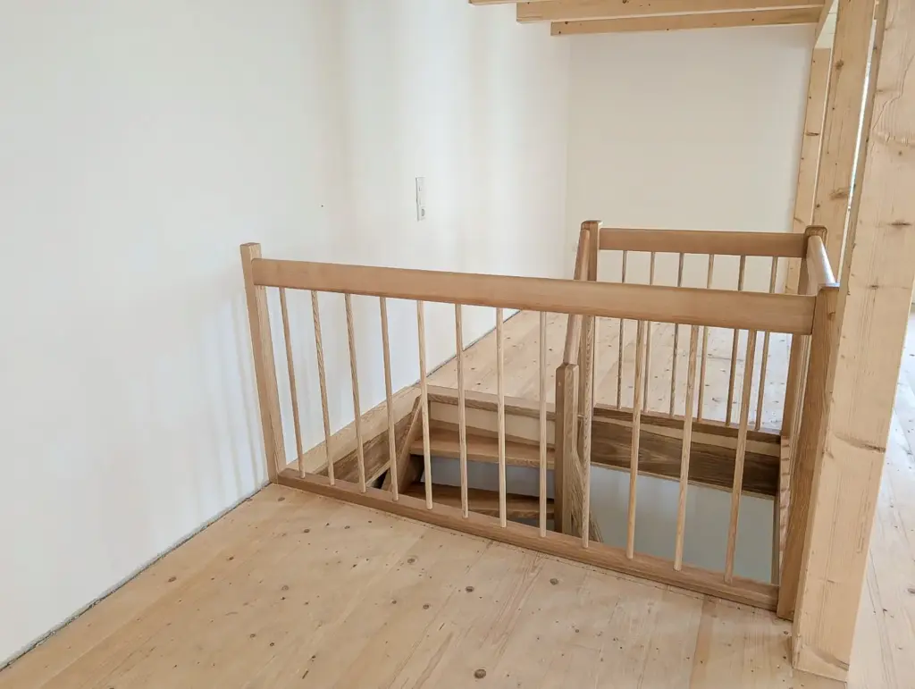Eine Holztreppe mit Sicherheitstor und Geländer führt von einer oberen Etage mit hellem Holzboden und schlichten weißen Wänden hinunter.