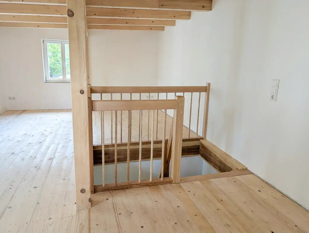 Ein heller, unmöblierter Dachboden mit hellen Holzböden und Balken, der über eine kleine Treppe mit einem Sicherheitsgeländer aus Holz zu einer unteren Ebene führt. Ein Fenster sorgt für natürliches Licht.