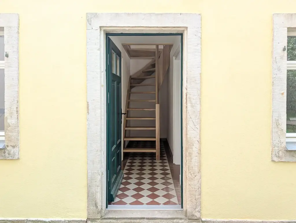 Eine geöffnete türkisfarbene Tür in einer gelben Wand gibt den Blick frei auf einen schmalen Flur mit einem karierten Fliesenboden und einer Holztreppe, die nach oben führt.