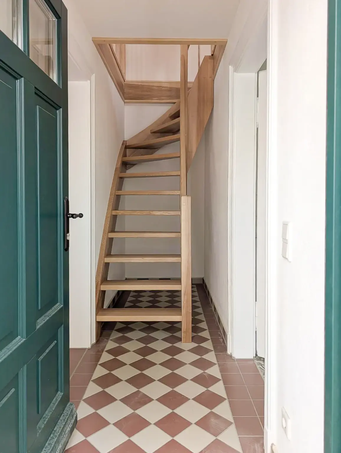 Eine Holztreppe mit offenen Setzstufen führt von einem schmalen Flur nach oben. Der Flur hat gemusterte Bodenfliesen in Braun und Weiß, weiße Wände und eine offene dunkelgrüne Tür auf der linken Seite.