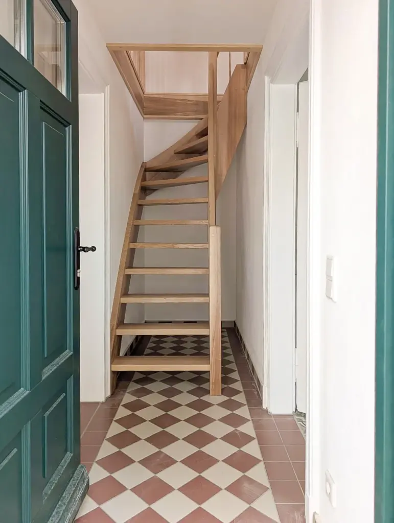 Eine Holztreppe mit offenen Setzstufen führt von einem schmalen Flur nach oben. Der Flur hat gemusterte Bodenfliesen in Braun und Weiß, weiße Wände und eine offene dunkelgrüne Tür auf der linken Seite.