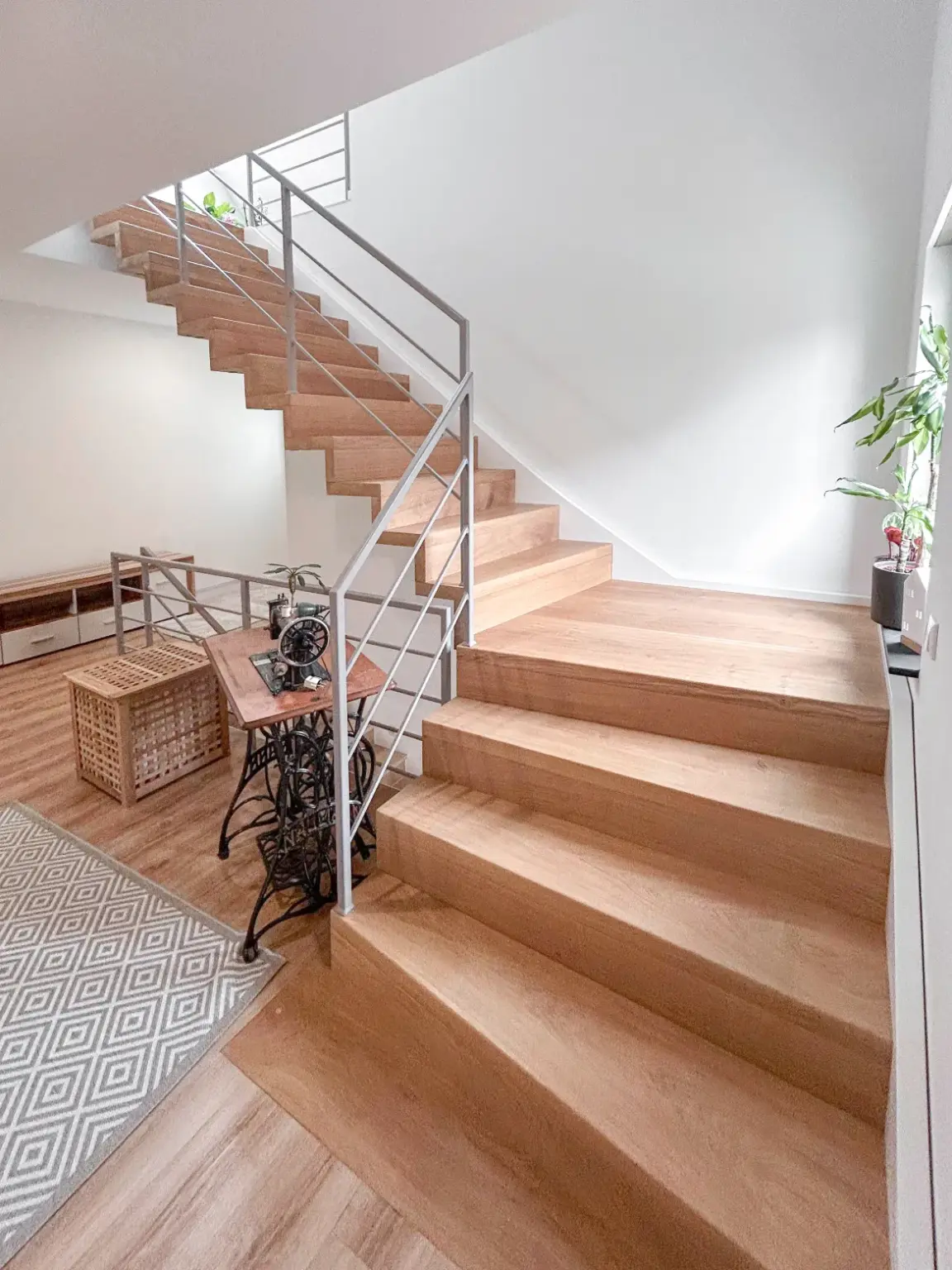 Eine moderne Innentreppe mit Holzstufen und Metallgeländer, umgeben von hellen Wänden. Auf dem Sockel steht ein kleiner Tisch mit einer Pflanze und einer alten Nähmaschine, daneben ein geometrisch gemusterter Teppich.