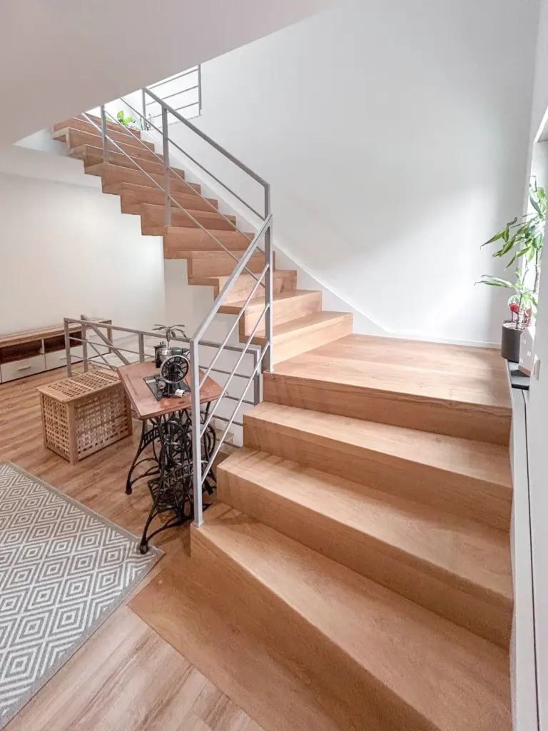 Eine moderne Innentreppe mit Holzstufen und Metallgeländer, umgeben von hellen Wänden. Auf dem Sockel steht ein kleiner Tisch mit einer Pflanze und einer alten Nähmaschine, daneben ein geometrisch gemusterter Teppich.