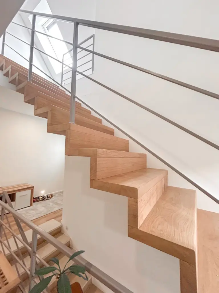 Eine moderne, minimalistische Holztreppe mit scharfen, kantigen Stufen und einem eleganten Metallgeländer in einem hellen, weiß getäfelten Innenraum, der durch ein Fenster von oben mit natürlichem Licht beleuchtet wird.