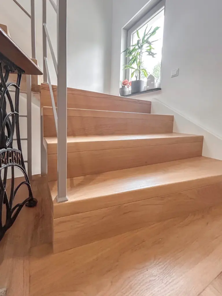 Eine Holztreppe mit vier Stufen führt zu einem Treppenabsatz in der Nähe eines Fensters; auf der Fensterbank stehen eine Topfpflanze und kleine Dekorationsgegenstände, und links der Treppe befindet sich ein Metallgeländer.