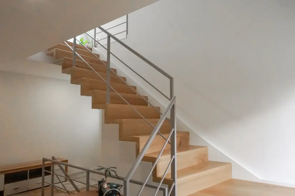 Moderne Holztreppe mit schlichtem Metallgeländer in einem hellen, minimalistischen Interieur. Am oberen Ende der Treppe steht eine kleine Pflanze und unten ist ein Möbelstück teilweise sichtbar. Weiße Wände und natürliches Licht erfüllen den Raum.