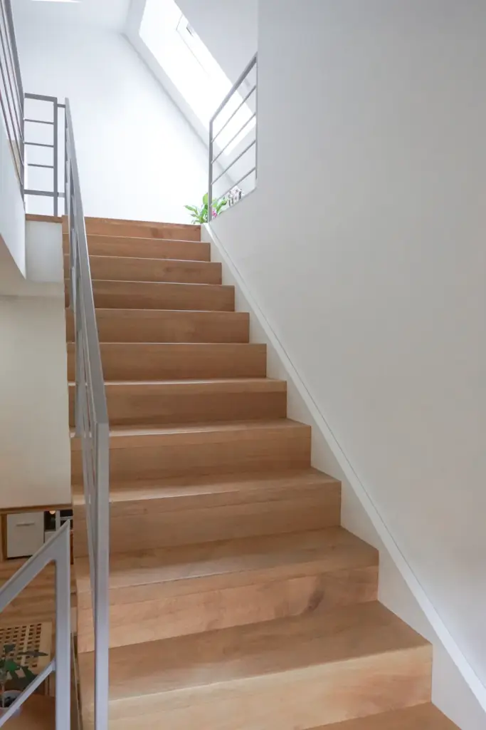 Eine moderne Innentreppe mit breiten Holzstufen und einem eleganten Metallgeländer führt zu einem Oberlicht hinauf, das die minimalistischen weißen Wände mit natürlichem Licht erhellt. Eine kleine Grünpflanze ist am oberen Ende der Treppe zu sehen.