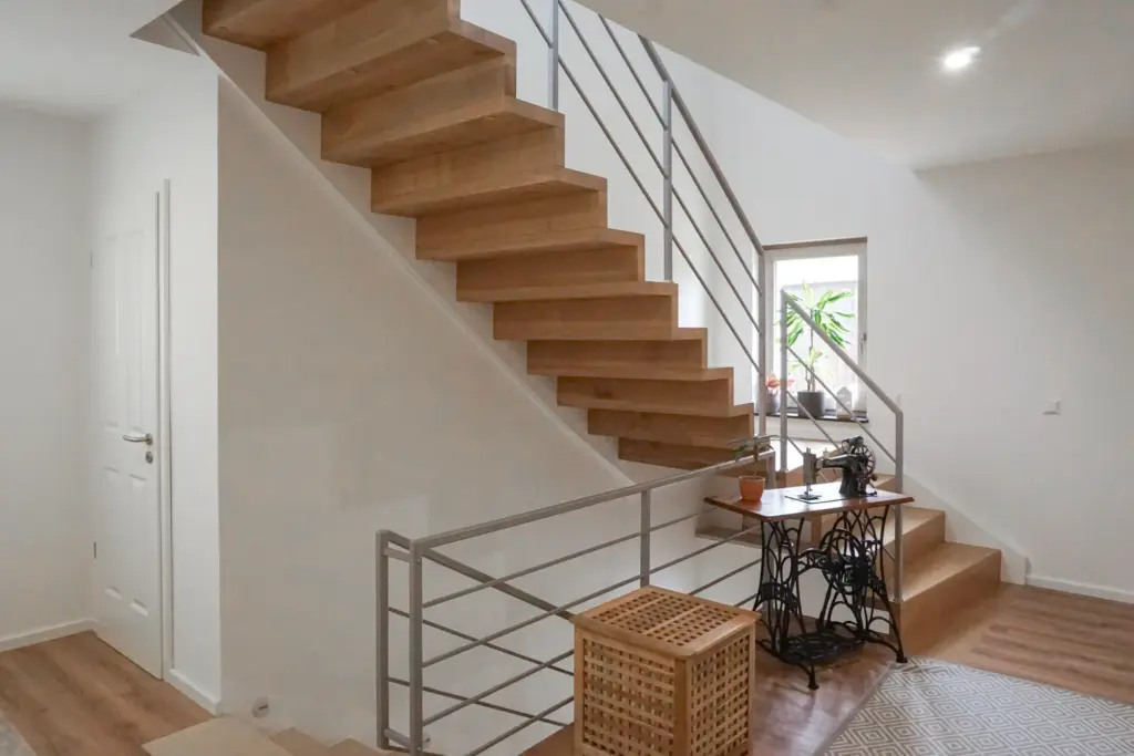 Ein modernes Interieur mit Holztreppen, weißen Wänden, einem Metallgeländer, einem alten Nähmaschinentisch, einem geflochtenen Korb und einem Fenster mit Topfpflanzen, das natürliches Licht hereinlässt.
