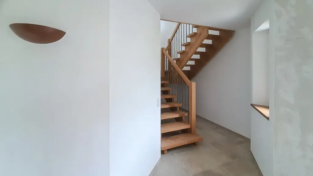 Moderne Holztreppe mit offenen Setzstufen und Metallgeländer in einem hellen, minimalistischen Innenraum mit weißen Wänden und großen Fliesenböden. Natürliches Licht fällt durch ein Fenster auf der rechten Seite ein.