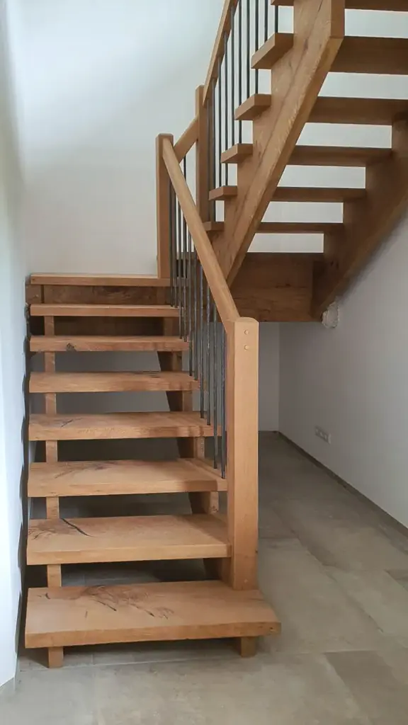 Eine Holztreppe mit offenen Setzstufen und Metallgeländer führt in ein Obergeschoss in einem modernen, minimalistischen Interieur mit hellen Fliesenböden und weißen Wänden.