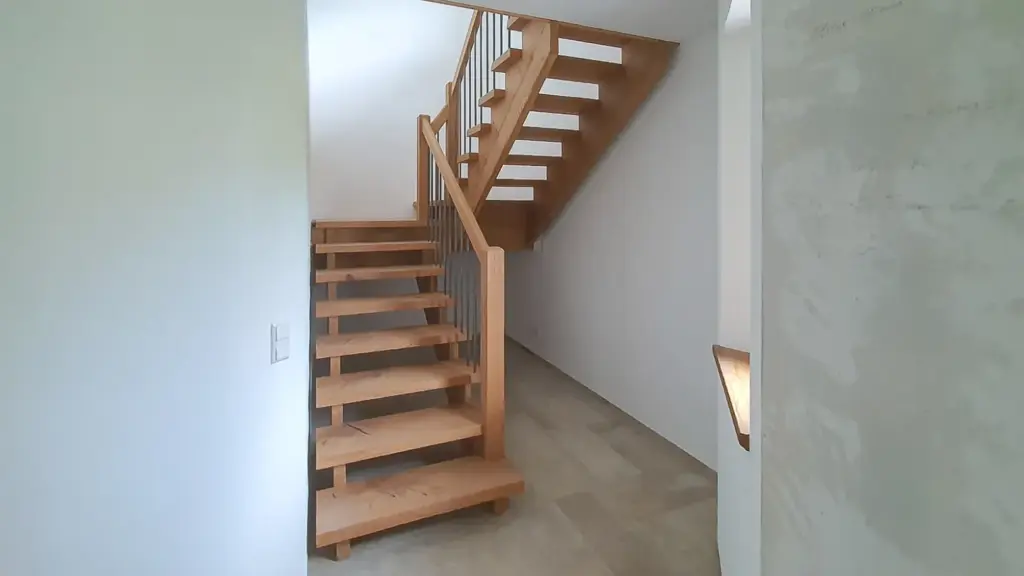 Eine moderne Holztreppe mit offenen Setzstufen und Metallgeländer befindet sich in einem hellen, minimalistischen Innenraum mit weißen Wänden und einem hellen Boden. Die Treppe dreht sich an einem Podest und führt weiter nach oben.