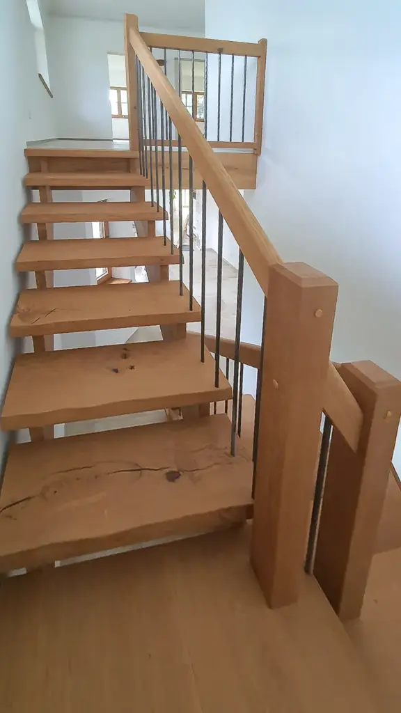 Eine moderne Holztreppe mit offenen Setzstufen, dicken Bohlen und schwarzen Metallgeländern führt zu einem Treppenabsatz in einem hellen, weiß getäfelten Innenraum.