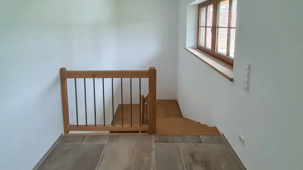 Eine Holztreppe mit einem Sicherheitstor am oberen Ende führt von einem Raum mit gefliesten Böden, weißen Wänden und einem Fenster mit Holzverkleidung hinunter, das Tageslicht hereinlässt.
