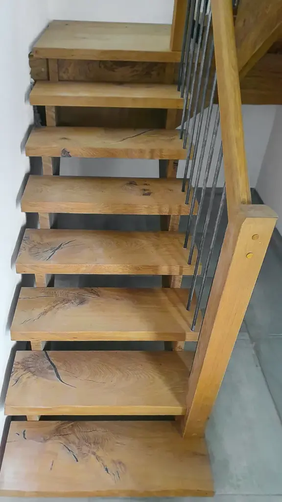 Eine Holztreppe mit breiten, rustikal anmutenden Stufen und einem Geländer mit vertikalen Metallstäben vor einem gefliesten Boden und einer weißen Wand.