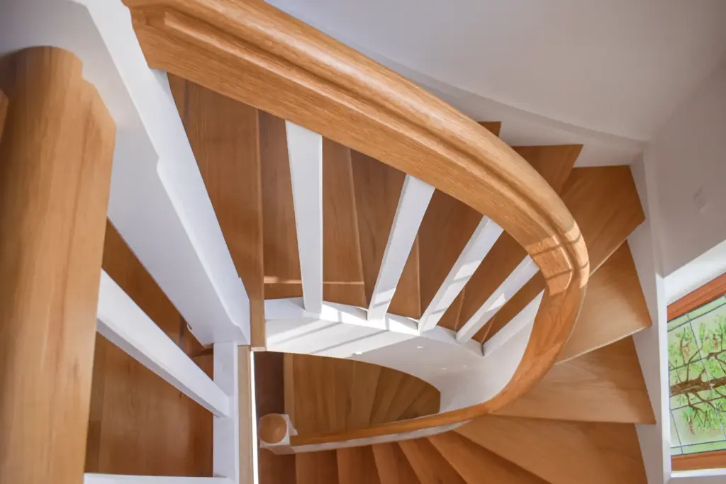 Blick von oben auf eine moderne Holzspindeltreppe mit weißen Geländerstäben und einem geschwungenen Handlauf, die in einem hellen, gut beleuchteten Innenraum sanft und kreisförmig nach unten führt.