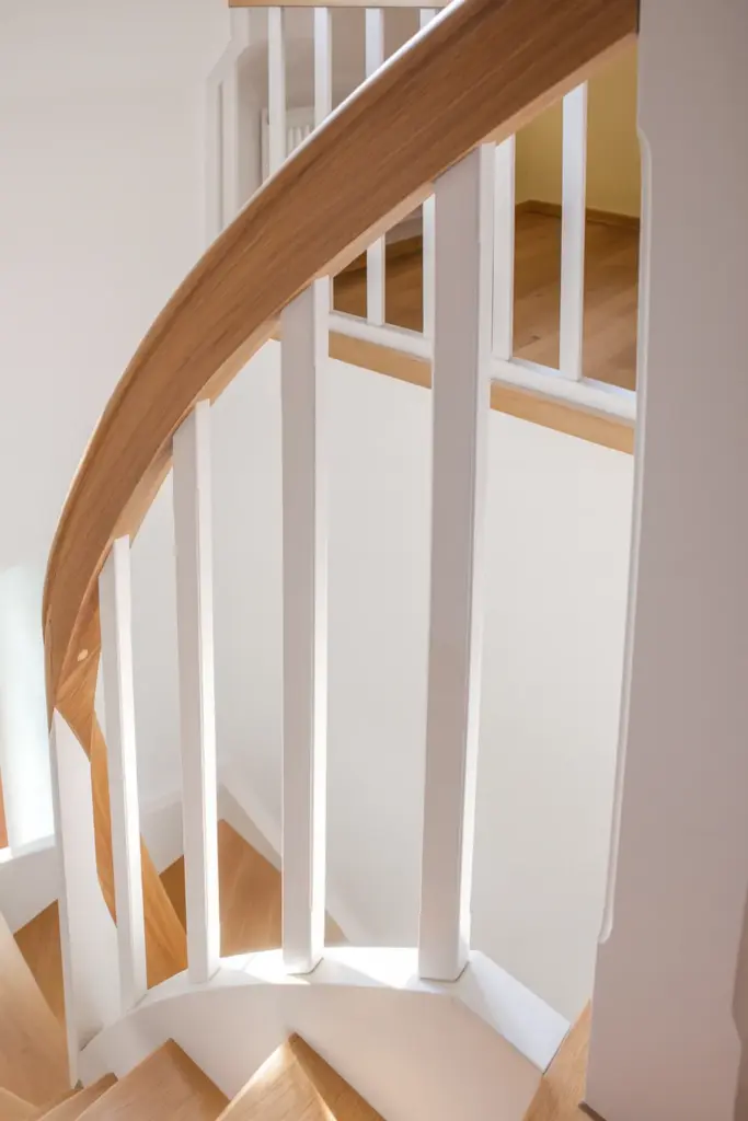 Geschwungene Holztreppe mit weißem vertikalen Geländer, hellen Holzstufen und passendem Handlauf, beleuchtet von natürlichem Licht, das aus einem nahe gelegenen Fenster kommt.
