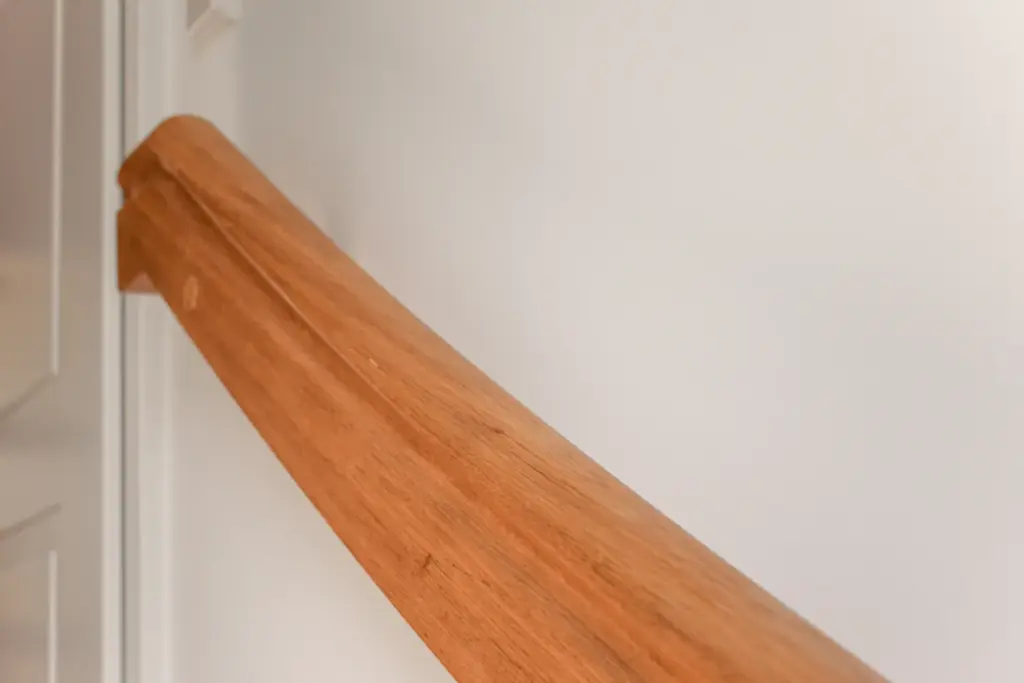 Nahaufnahme eines hölzernen Handlaufs, der an einer weißen Wand befestigt ist, wobei im Hintergrund ein Teil einer weißen Tür zu sehen ist.