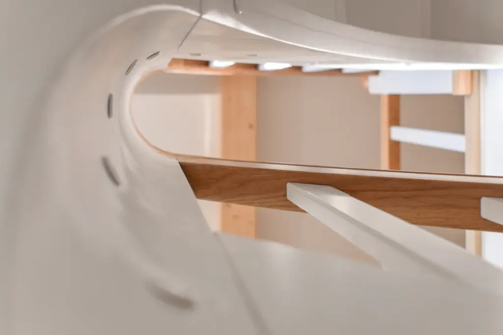 Nahaufnahme einer modernen Wendeltreppe mit weißen Stufen und einem Handlauf aus hellem Holz, die abstrakte Kurven und Linien in einem gut beleuchteten Innenraum erzeugt.