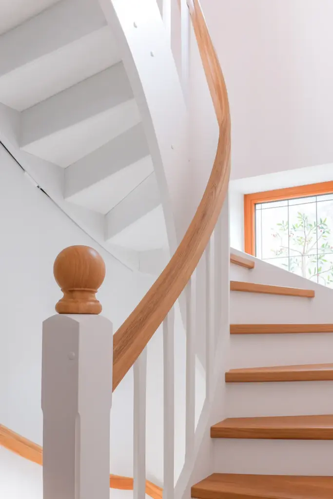 Eine moderne Wendeltreppe mit weißen Stufen und Geländern, mit Akzenten aus Naturholz am Handlauf und an den Treppenstufen, neben einem sonnenbeschienenen Fenster mit Holzrahmen.