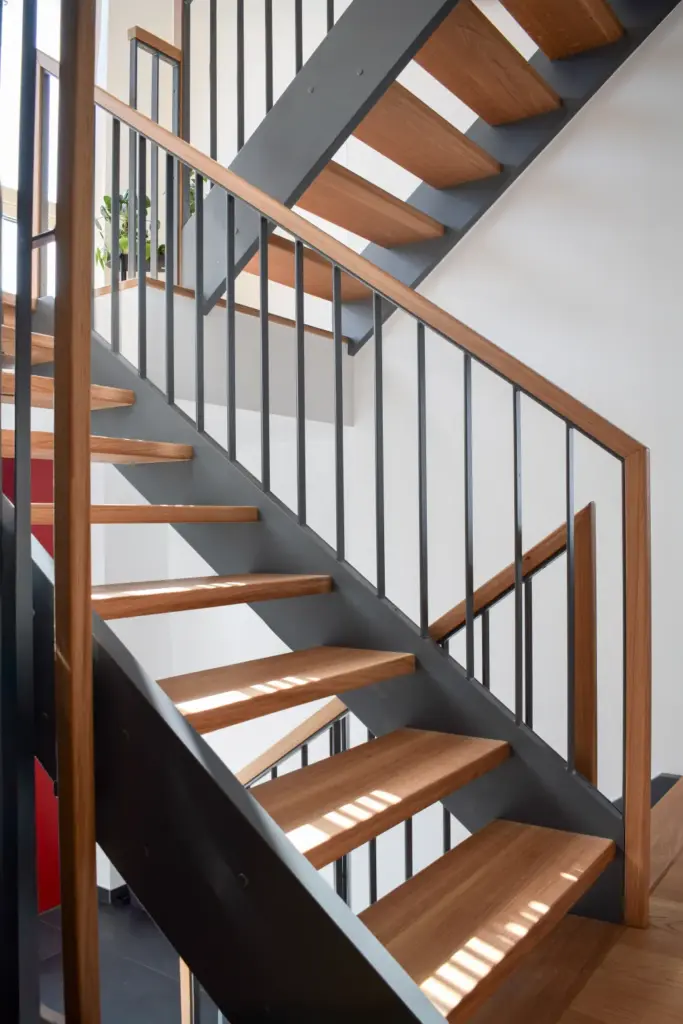 Eine moderne Treppe mit Holzstufen und schwarzem Metallgeländer, die von Tageslicht erhellt wird, führt an einer weißen Wand entlang nach oben; auf dem Treppenabsatz darüber ist eine Topfpflanze zu sehen.
