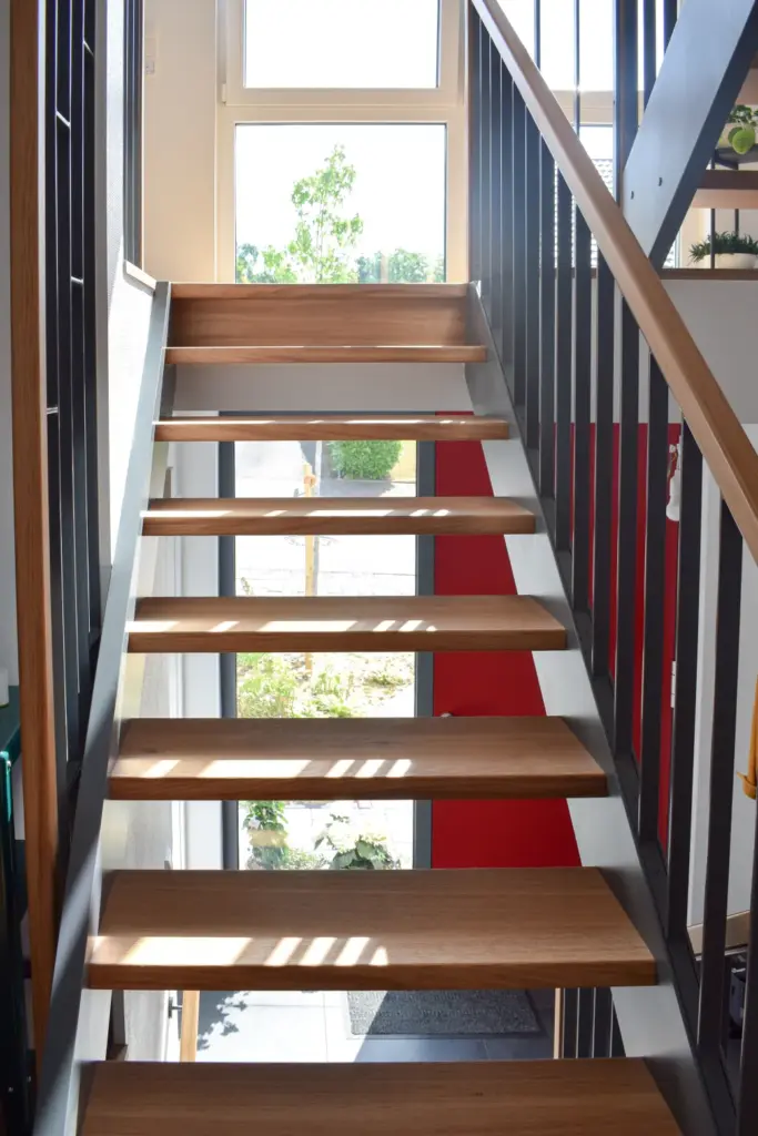Moderne Innentreppe mit Holzstufen, schwarzem Metallgeländer und Sonnenlicht, das durch große Fenster fällt und Schatten auf der Treppe und dem Boden erzeugt. Draußen sind ein Baum und Grünflächen zu sehen.
