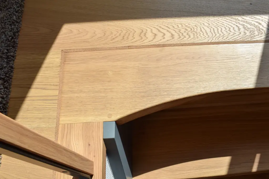 Das Sonnenlicht wirft Schatten auf den hellen Holzboden und den dazu passenden Holztisch mit einem grauen Metallbein, wodurch geometrische Muster entstehen. In der oberen linken Ecke ist ein Teil eines strukturierten dunklen Teppichs zu sehen.
