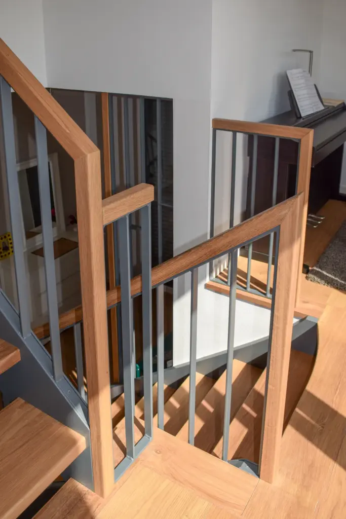Moderne Holztreppe mit grauem Metallgeländer und quadratischen Pfosten, beleuchtet von natürlichem Sonnenlicht, neben einer Ecke mit einem Klavier und einem Teppich auf einem Holzboden.