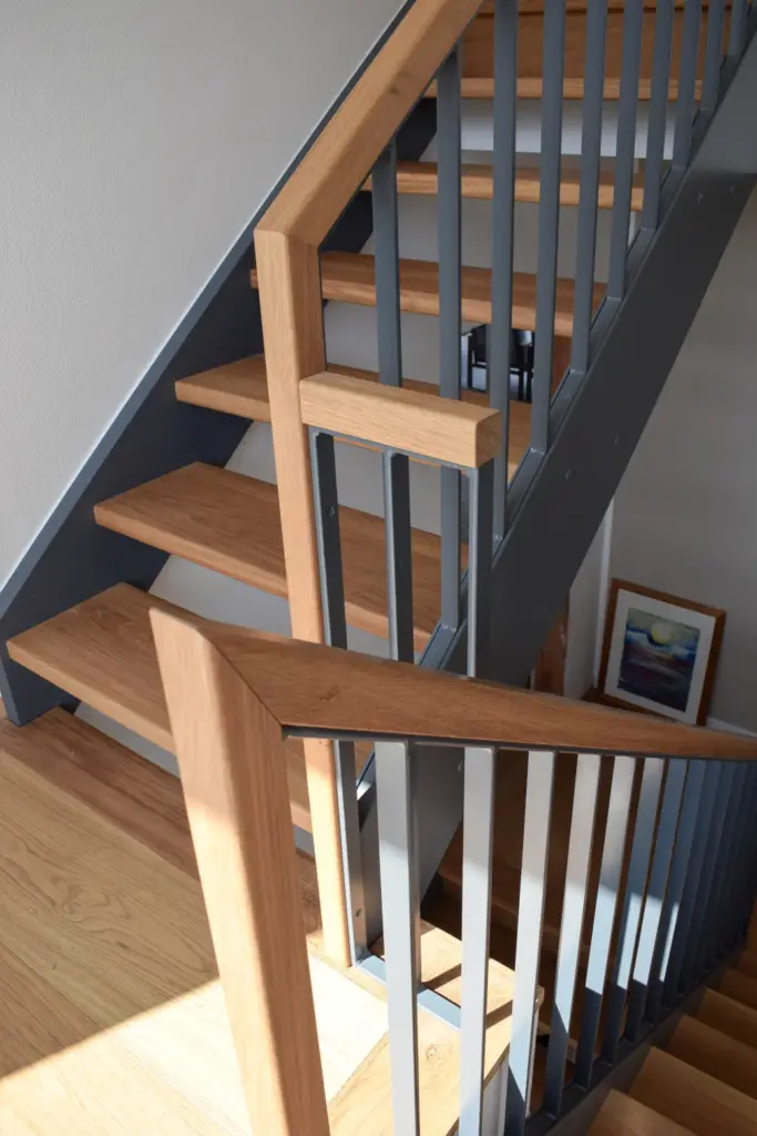 Eine moderne Treppe mit hölzernen Stufen und Handläufen, grauen Metallgeländern und einem Gemälde, das an der Wand im unteren Stockwerk hängt. Das Sonnenlicht wirft Schatten auf die Treppe und die Wände.