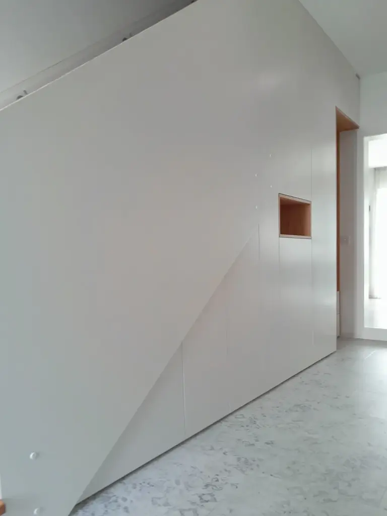 Minimalistisches weißes Treppenhaus mit geometrischen Linien und Einbauschränken mit einem kleinen offenen Holzregal; der Raum wirkt sauber und modern mit einem hellen Marmorboden.