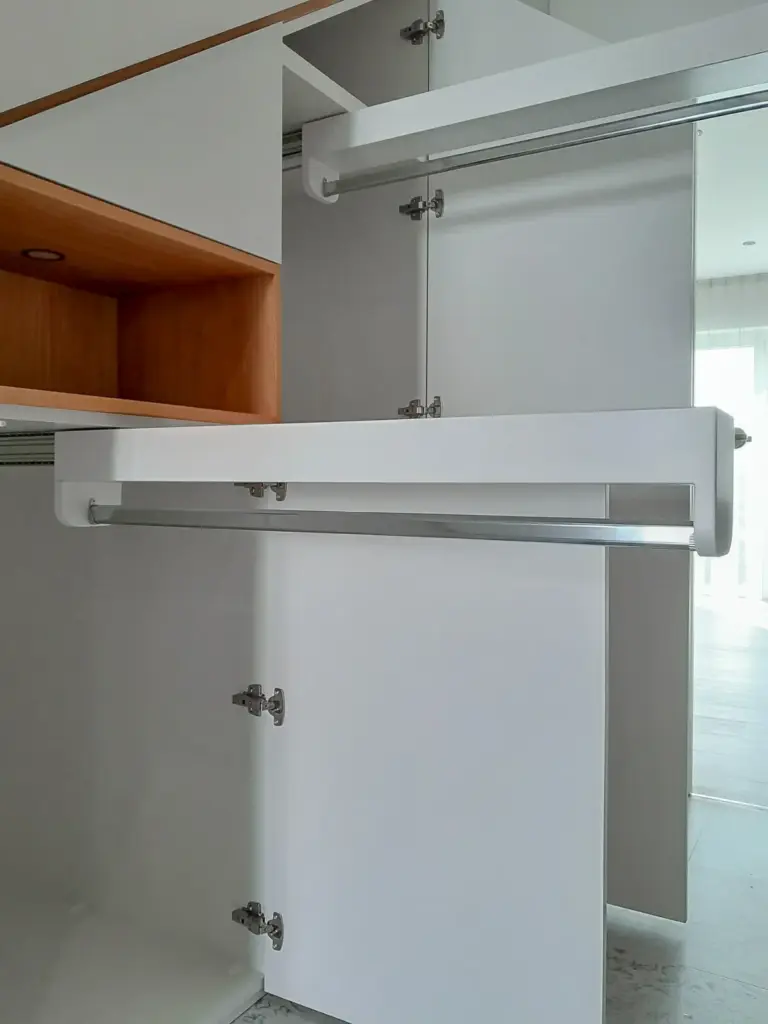 Offener weißer Kleiderschrank mit sichtbaren Metallstangen, Einlegeböden und Scharnieren, mit natürlichem Licht aus einem Fenster im Hintergrund.