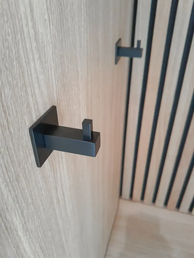 Zwei moderne, schwarze Metallhaken sind übereinander an einer hellen Holzwand mit vertikalen Rillen angebracht. Die Szene hat eine minimalistische und zeitgenössische Designästhetik.