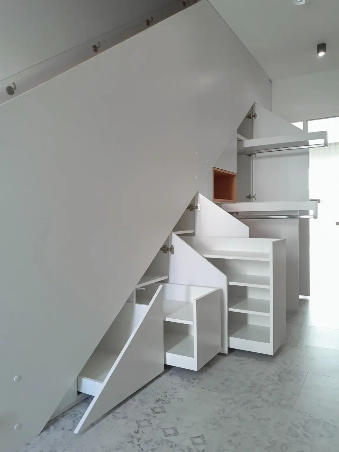 Weißer Stauraum unter der Treppe mit mehreren offenen Regalen, ausziehbaren Schubladen und Schränken. Das klare, moderne Design maximiert den Platz unter der Treppe in einem hellen, minimalistischen Raum.