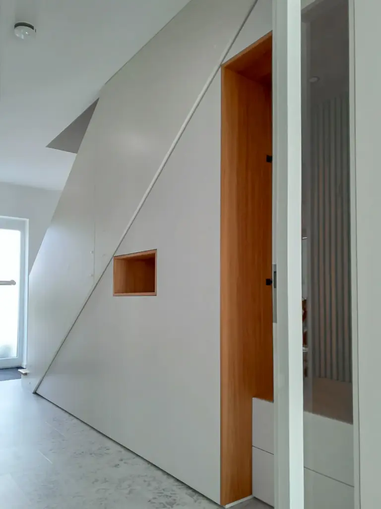 Ein moderner Innenflur mit einer schrägen weißen Wand mit eingebauter Holznische und Regal sowie einer teilweise offenen Tür, die eine vertikale Lamellenverkleidung im Inneren offenbart. Der Raum ist minimalistisch und hell.