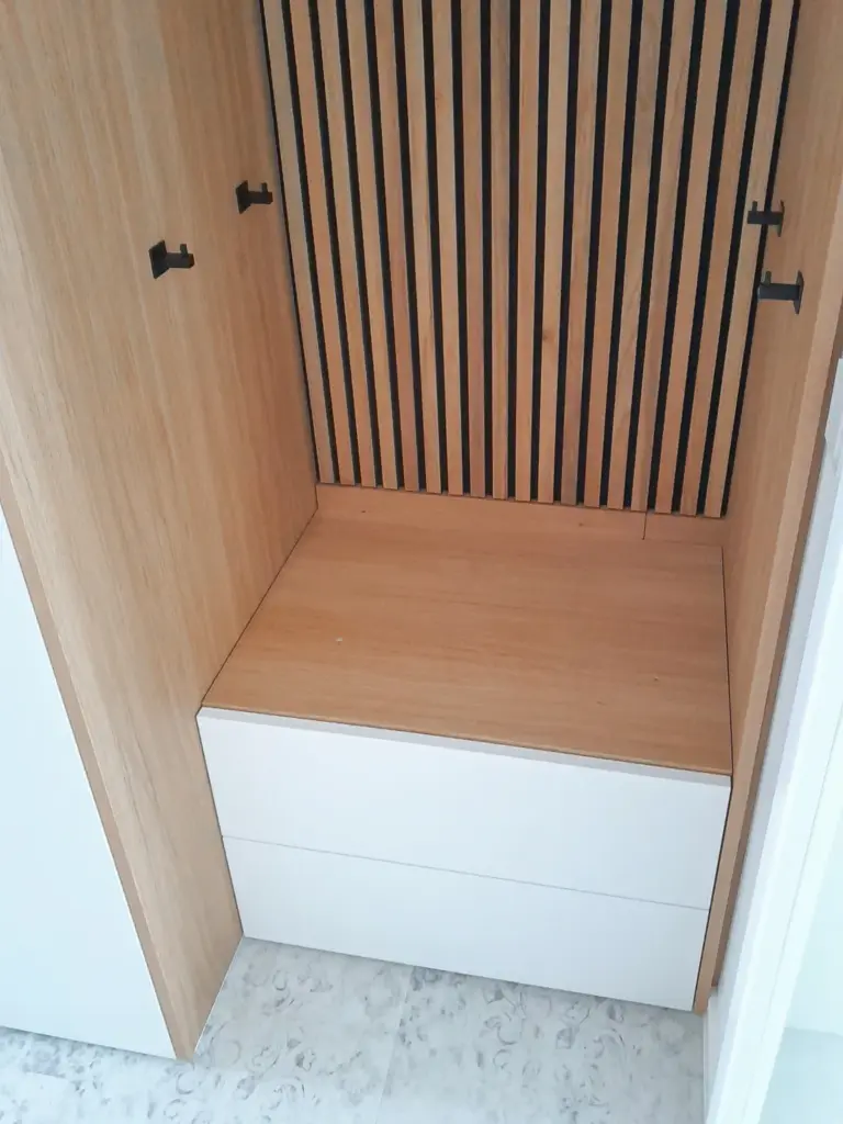 Ein hölzerner Schrank mit offenen Türen gibt den Blick auf eine Bank mit einer hellen Holzplatte und zwei weißen Schubladen darunter frei. An der Rückwand befinden sich vertikale schwarze Latten vor einem hölzernen Hintergrund. Der Boden ist mit hellen Fliesen ausgelegt.