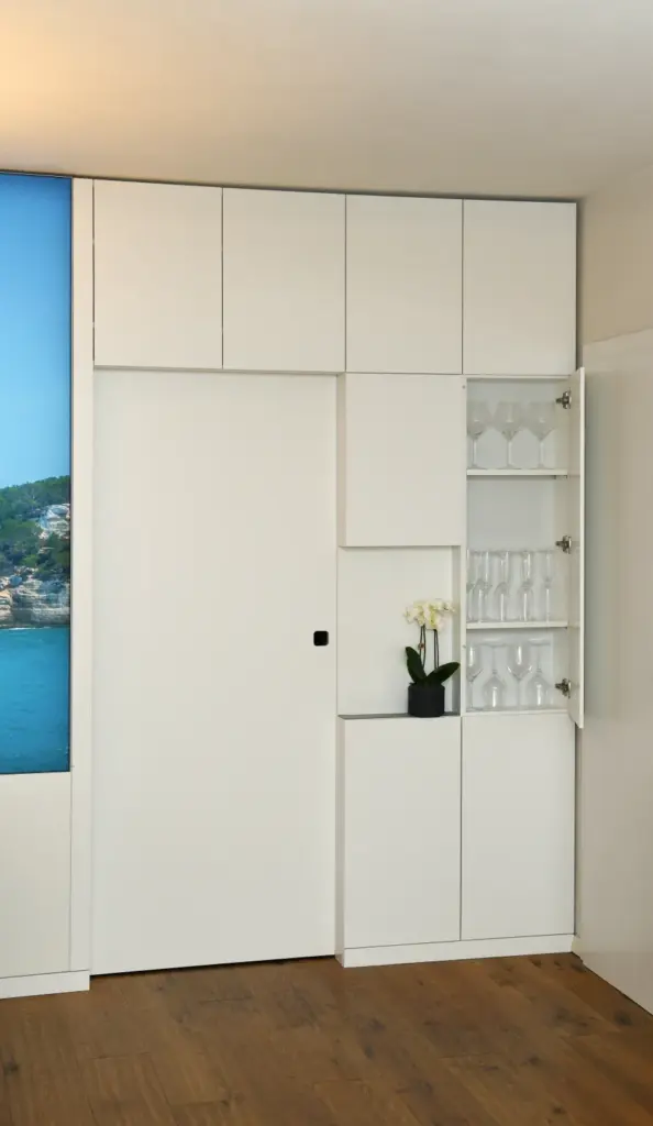 Ein moderner weißer Einbauschrank mit offenen Regalen, in denen Glaswaren und eine kleine Topforchidee stehen, neben einer geschlossenen Schiebetür auf einem Holzboden.