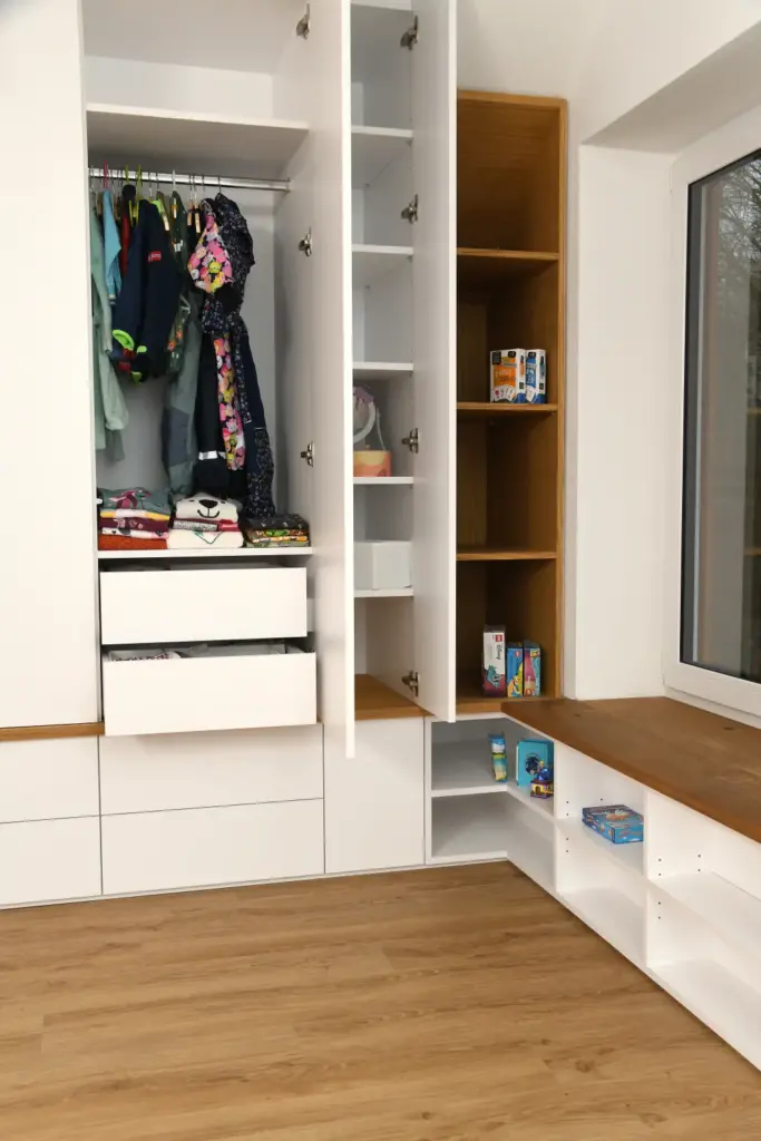 Offener weißer Kleiderschrank mit Kleidung, offenen Schubladen und Regalen mit Spielzeug und Büchern. Eine Holzbank mit Stauraum steht unter einem Fenster, alles auf einem hellen Holzboden.