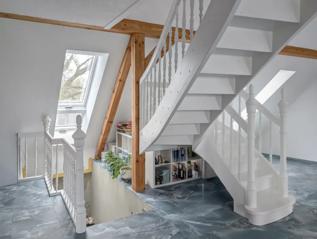 Ein modernes Loft mit weißer Holztreppe, marmoriertem Boden, Dachfenster, Holzbalken, weißem Geländer und einem in die Treppe eingebauten Bücherregal. Natürliches Licht erhellt den Raum.