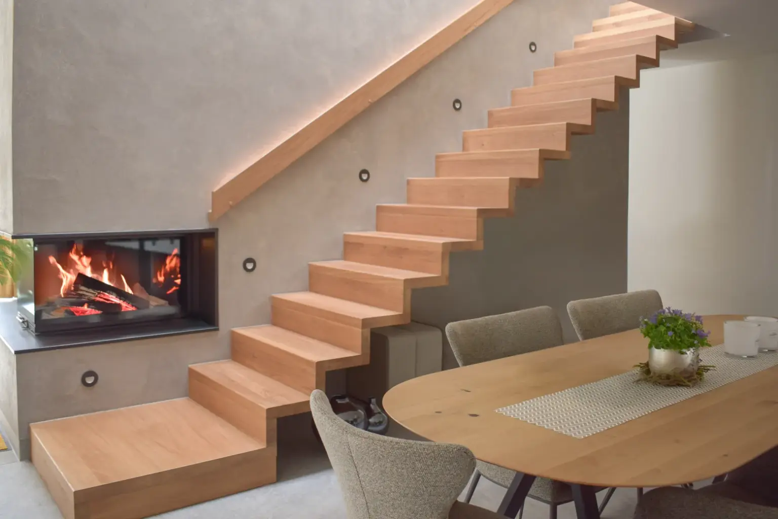 Ein modernes Interieur mit einer schwebenden Holztreppe, einem in die Wand unter der Treppe eingebauten Kamin und einem hölzernen Esstisch mit Stühlen und einem Pflanzenaufsatz.