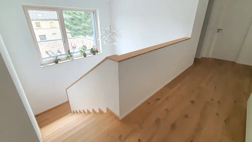Ein moderner, heller Flur mit hellem Holzfußboden, weißen Wänden, einer Treppe mit Holzstufen und einem Fenster mit mehreren Topfpflanzen auf der Fensterbank. In der Nähe des Fensters hängt ein dekorativer weißer Kronleuchter.