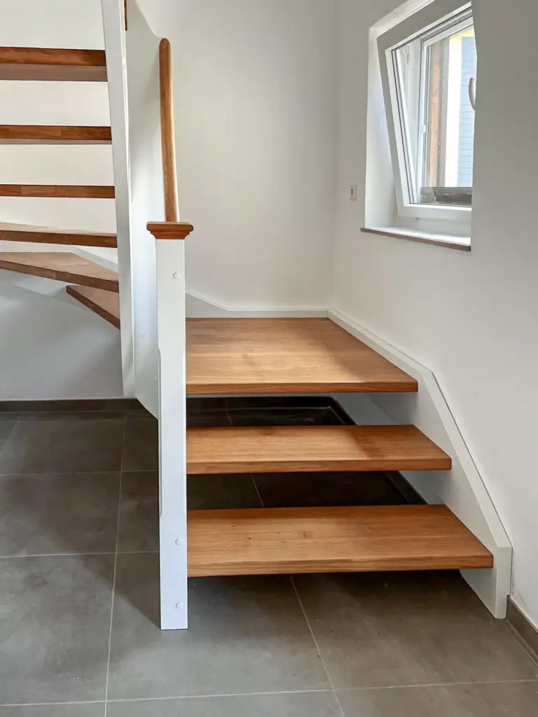Eine moderne Holztreppe mit drei schwimmenden Stufen und einem weißen Geländer steht in einem hellen Raum mit grauem Fliesenboden und einem Fenster an der rechten Wand.
