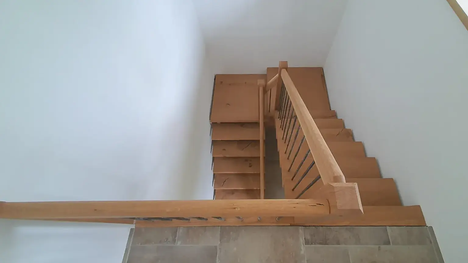 Eine Holztreppe mit einem einfachen Handlauf führt von einem gefliesten Treppenabsatz hinunter, der von schlichten weißen Wänden umgeben ist.