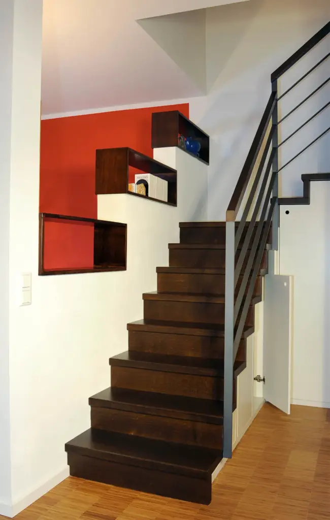 Eine moderne Holztreppe mit dunklen Stufen und einem Metallgeländer, mit offenen rechteckigen Regalen an einer rot-weißen Wand und einer kleinen Tür unterhalb der Treppe auf einem hellen Holzboden.