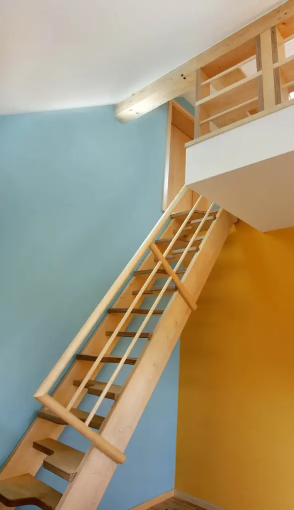 Eine Holztreppe mit Geländer führt hinauf zu einem Dachboden in einem hell erleuchteten Raum mit blauen und gelben Wänden. Der Dachboden hat ein Holzgeländer und eine oben sichtbare Tür.