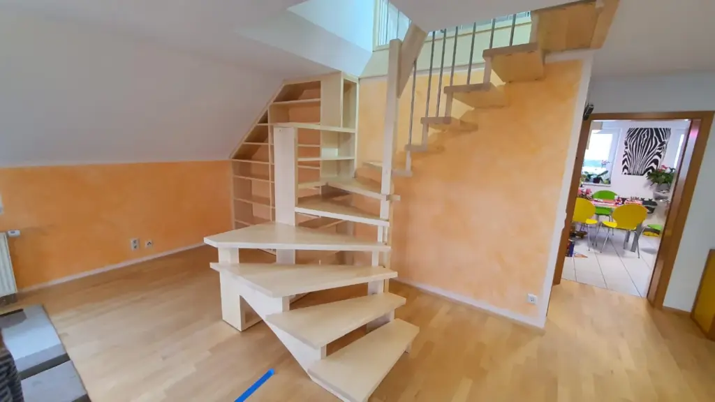 Eine moderne, helle Holztreppe mit offenen Setzstufen und darunter liegenden Einbauregalen vor orangefarbenen Wänden in einem hellen Raum. Die Treppe führt zu einem oberen Stockwerk mit Metallgeländer.