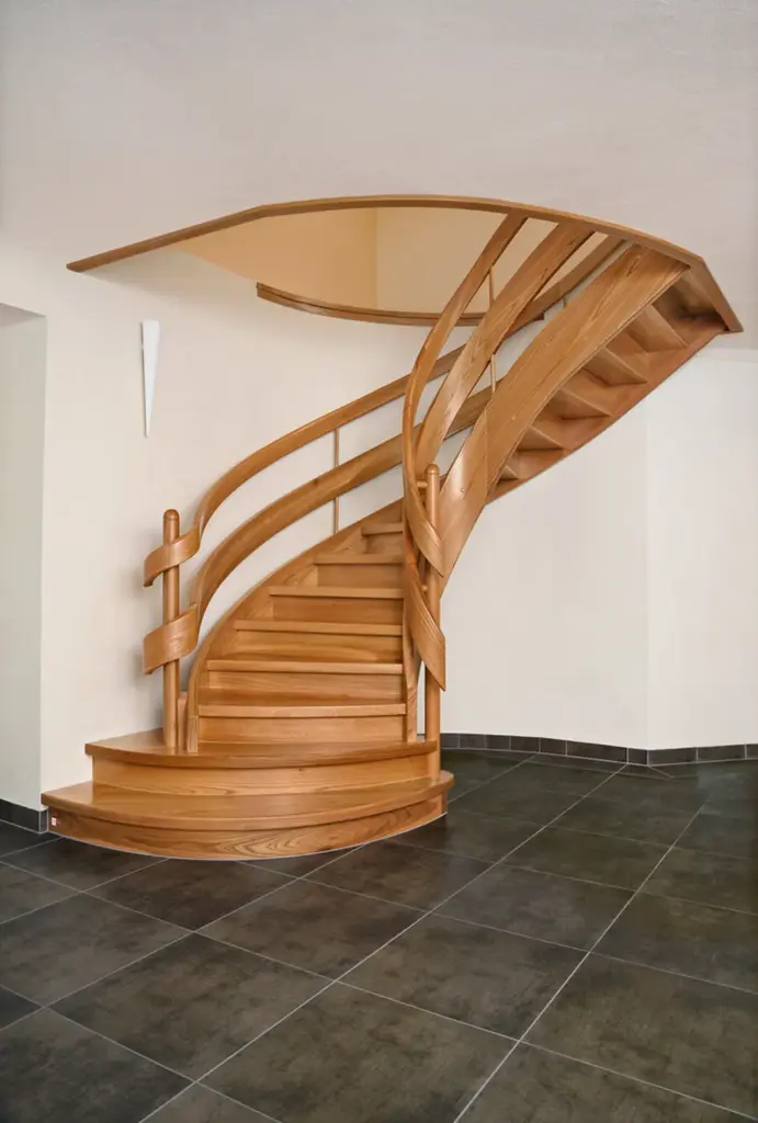 Eine moderne Holzspindeltreppe mit glattem, geschwungenem Geländer steht auf einem dunklen Fliesenboden vor cremefarbenen Wänden. Die Treppe führt nach oben und verschwindet teilweise in der Decke.