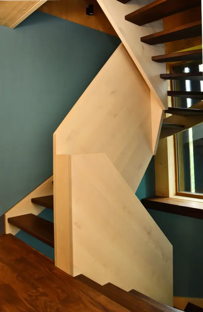 Eine moderne Holztreppe mit kantigen, hellen Holzpaneelen und dunklen Holzstufen, die vor einer blaugrünen Wand und neben einem Fenster steht, das natürliches Licht hereinlässt.
